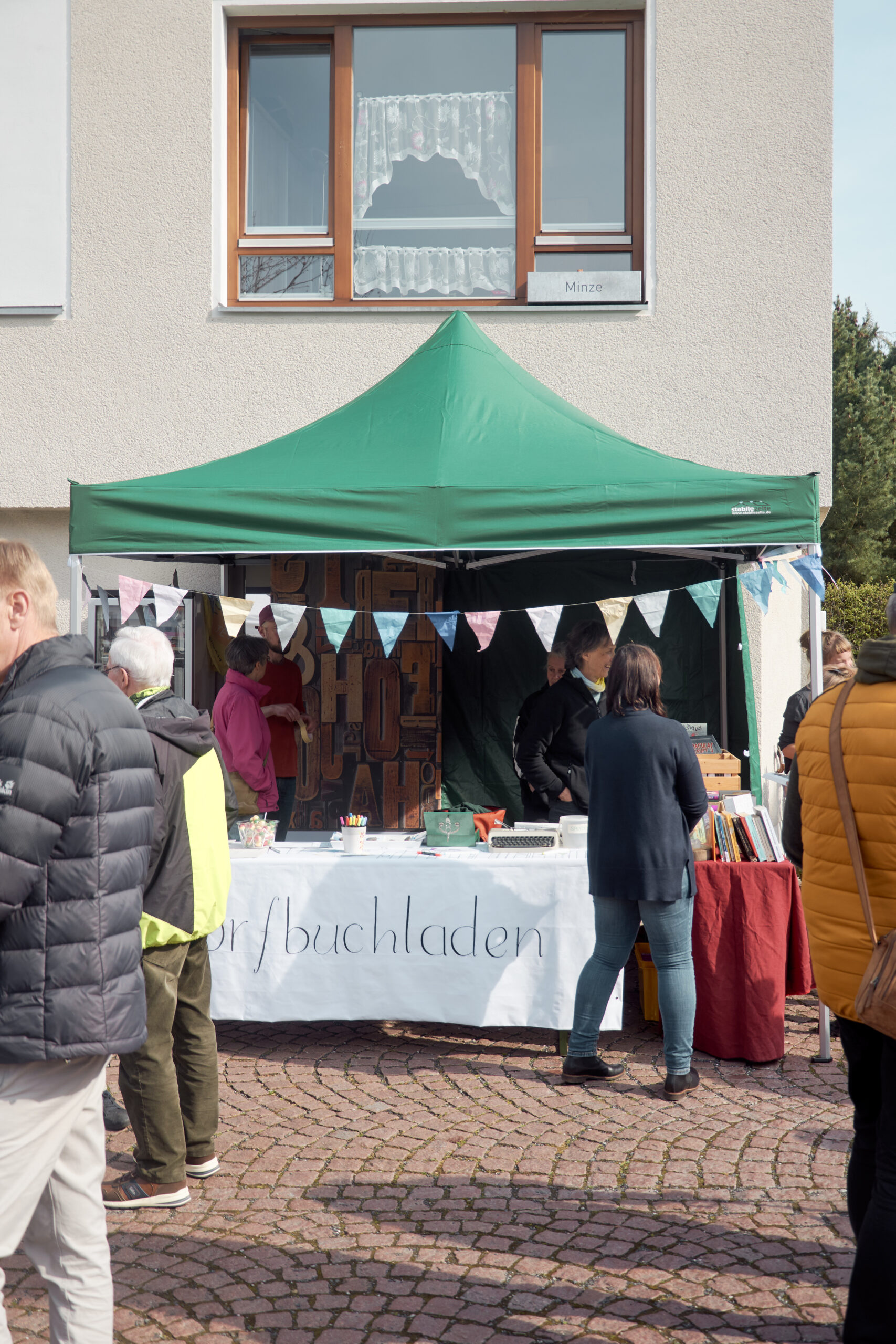 zeigt einen grünen Pavillon mit weißem Banner, wo "Dorfbuchladen" draufsteht.